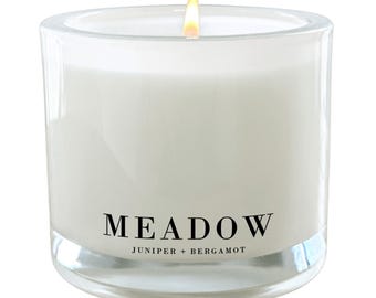 Puede incluir: Un tarro de vela de vidrio transparente con una vela blanca en su interior. La vela está encendida y la llama es visible. El tarro tiene una etiqueta negra que dice "MEADOW JUNIPER + BERGAMOT".