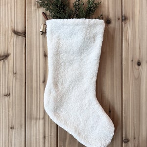 以下が含まれることがあります： 白いふわふわのクリスマスストッキングに、金色のベルがぶら下がっています。ストッキングには、黒い文字で「EVIE」と書いてあります。