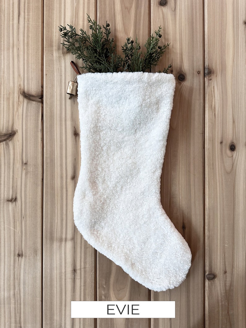 以下が含まれることがあります： 白いふわふわのクリスマスストッキングに、金色のベルがぶら下がっています。ストッキングには、黒い文字で「EVIE」と書いてあります。