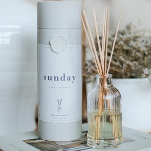 May include: A reed oil diffuser set with a clear glass bottle and wooden reeds. The bottle is filled with a light yellow liquid. A cylindrical container with the word "sunday" in blue lettering is next to the bottle. The container has a white wax seal.