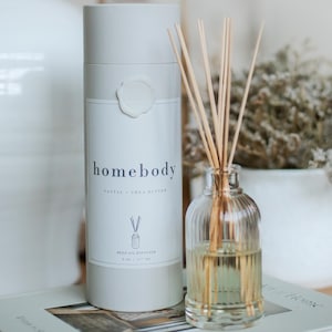 May include: A reed oil diffuser with a clear glass bottle and wooden reeds. The bottle is filled with a light yellow liquid. A cylindrical container with the text "homebody" and "Santal + Shea Butter" is next to the diffuser.