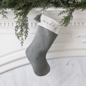 May include: A gray Christmas stocking with a white cuff, hanging from a garland. The cuff is embroidered with the name "molly" in black script. A gray tassel hangs from the top of the stocking.