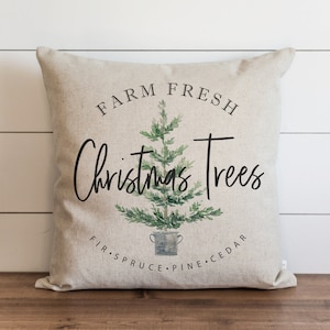 Op de afbeelding: Een beige kussen met de tekst "Farm Fresh Christmas Trees" en een aquarel-illustratie van een kerstboom in een emmer. De tekst "Fir Spruce Pine Cedar" staat onder de boom.
