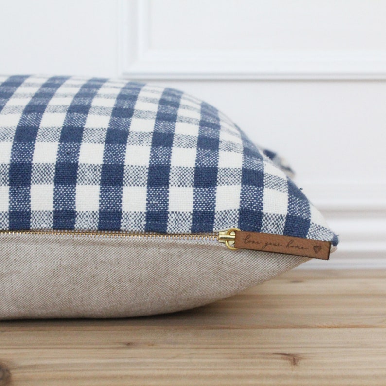 K&ouml;nnte beinhalten: Ein blaues und wei&szlig;es Gingham-Kissen mit Rei&szlig;verschluss und einem braunen Lederanh&auml;nger mit der Aufschrift "Love your home" und einem Herz.