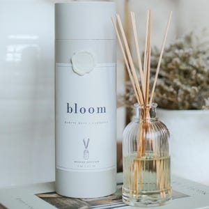 May include: A reed oil diffuser set with a clear glass bottle and a cylindrical container. The container is white with the word "bloom" in blue, and the scent is "Marine Moss + Gardenia". The set includes wooden reeds.
