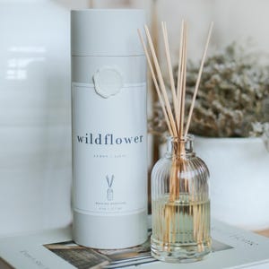 Puede incluir: Un juego de difusor de aceite de caña con una botella de vidrio transparente y cañas de madera. La botella está llena de un líquido amarillo claro. Un recipiente cilíndrico con la palabra "wildflower" está al lado de la botella. El aroma es limón y lila.
