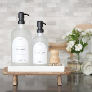 May include: Two frosted glass bottles with black pump tops and labels that read "No. 03 Dish Soap" and "No. 01 Hand Soap". The bottles are on a white rectangular tray with a wooden base. A wooden brush is in the tray.
