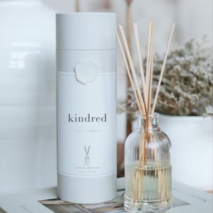 May include: A white cylindrical reed oil diffuser box with the word "kindred" printed on the front. A clear glass bottle with reeds is next to the box. The bottle contains a light yellow liquid. The text "Yuzu Freesia" and "Reed Oil Diffuser" are also visible.
