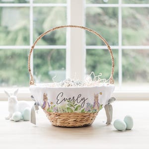 May include: A white Easter basket with a woven handle and a white fabric liner featuring a bunny illustration and the name "Everly". The basket is filled with shredded paper and blue Easter eggs.