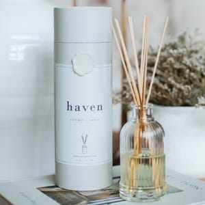 May include: A reed oil diffuser set with a cylindrical white container and a clear glass bottle. The container has the word "haven" in blue, with "saffron + amber" below it. The bottle contains a yellow liquid and wooden reeds.
