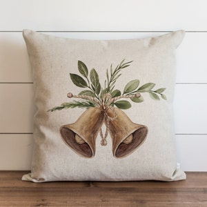 May include: A square beige pillow with a watercolor illustration of two bronze bells tied together with rope and adorned with green leaves. The pillow is on a wooden surface with a white wood-paneled background.
