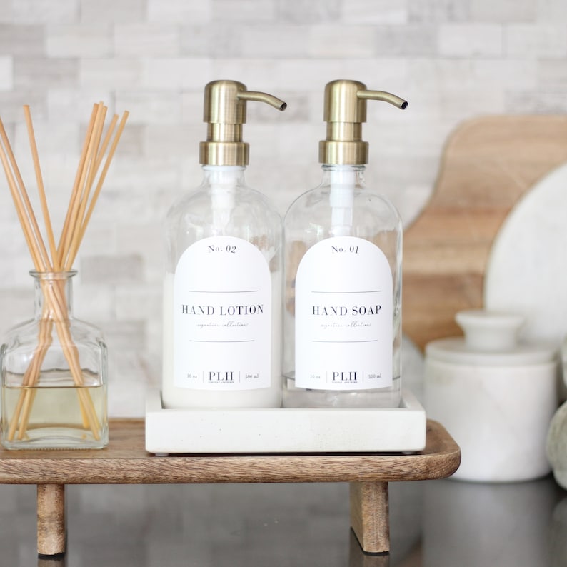 May include: Two glass bottles with gold pumps, one labeled "Hand Lotion" and the other "Hand Soap", sitting on a white tray on a wooden stand.