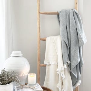 May include: A wooden ladder displays two blankets: one cream-colored with fringe and another gray with a chevron pattern. A white ceramic jar, a candle, and books are on a wooden side table, creating a cozy, neutral-toned home decor scene.