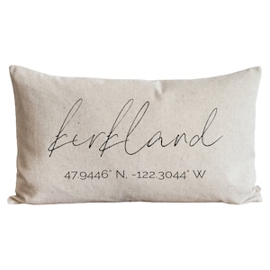 May include: A beige throw pillow with the word "Kirkland" written in cursive script and the coordinates "47.9446° N, -122.3044° W" printed below.