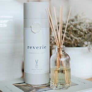 May include: A clear glass reed oil diffuser with wooden reeds and a white cylindrical container. The container has the word "reverie" printed on it, along with "Pomegranate + Rose" and "Reed Oil Diffuser". The diffuser is 6 oz / 177 ml.
