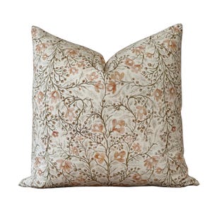 Puede incluir: Una almohada de color crema con un patrón floral en tonos marrones y naranjas. El patrón presenta ramas delicadas con pequeñas flores y hojas.