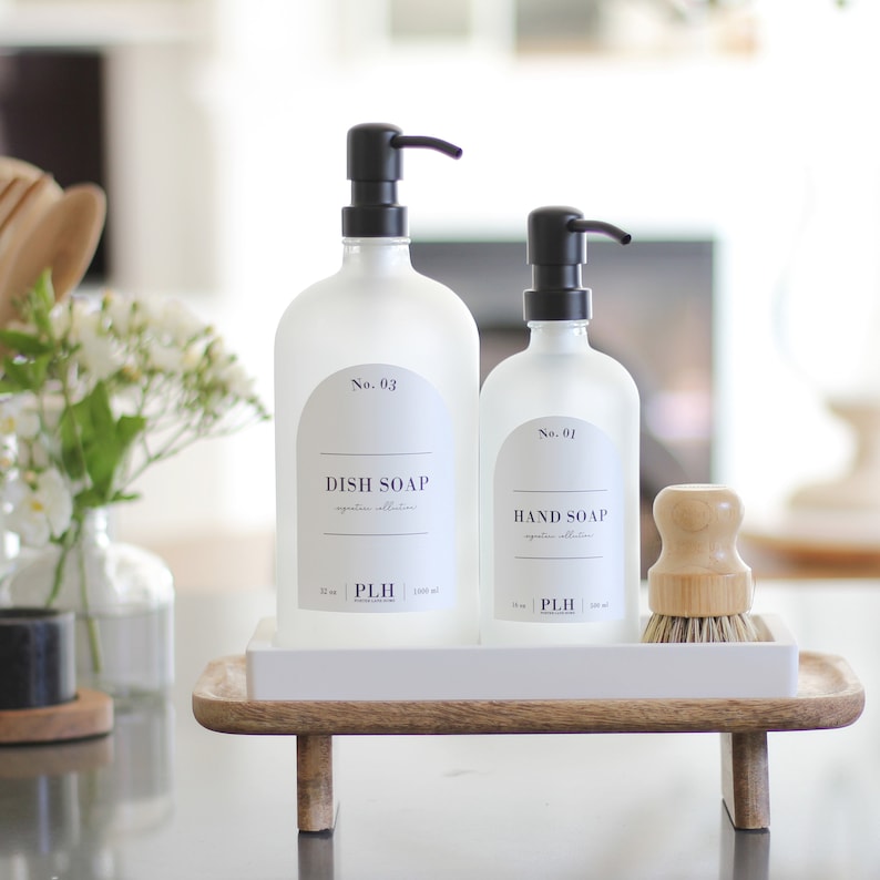 May include: Two frosted glass soap dispensers with black pumps on a white tray with wooden legs. The dispenser on the left is labeled "No. 03 DISH SOAP" and the dispenser on the right is labeled "No. 01 HAND SOAP". A small wooden brush is on the tray.