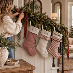 May include: Three Christmas stockings hanging from a staircase garland. The first is solid rust-colored fabric with a cream cuff labeled DAD. The second is cream fabric with a ruffled trim and cuff labeled MOM. The third is striped with a ruffled trim and cuff labeled PRESTON.