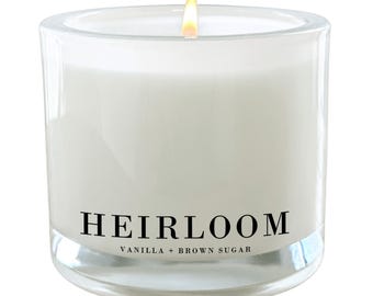 Puede incluir: Un tarro de vela de vidrio transparente con una vela blanca en su interior. La vela está encendida y la llama es visible. El tarro tiene una etiqueta negra que dice "HEIRLOOM VANILLA + BROWN SUGAR".