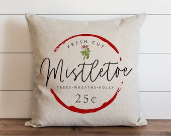 Puede incluir: Una almohada blanca con un círculo rojo y el texto "Fresh Cut Mistletoe Trees - Wreaths - Holly 25¢" en letras negras. El círculo tiene un efecto de salpicadura roja y una pequeña rama de muérdago verde con un lazo rojo.