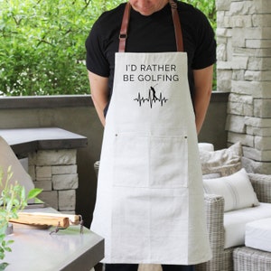 May include: White canvas apron with adjustable straps and a large pocket. The apron has a black graphic that reads "I'd rather be golfing" with a stylized heartbeat and a golfer silhouette.