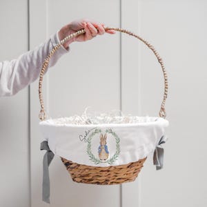 Puede incluir: Una cesta de mimbre tejida con un forro de tela blanca con una ilustración de Peter Rabbit y el nombre "Sadie". La cesta tiene un asa tejida y lazos de tela gris. La cesta está llena de papel blanco.