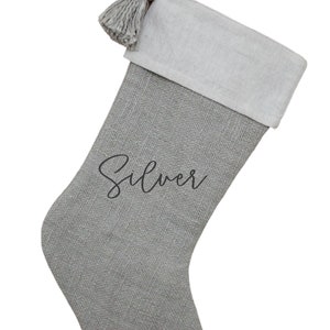 May include: A gray Christmas stocking with a white cuff and a matching tassel. The word "Silver" is written in cursive on the front. The stocking has a loop for hanging.