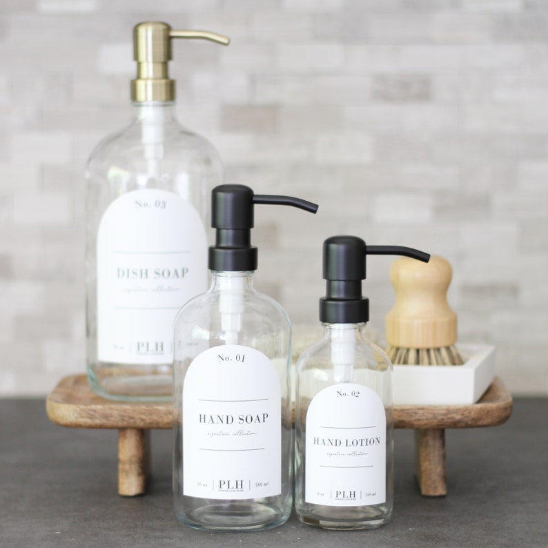 May include: Three glass bottles with black and gold pump tops. The bottles are labeled with white text on black labels. The labels read "No. 03 Dish Soap", "No. 01 Hand Soap", and "No. 02 Hand Lotion".