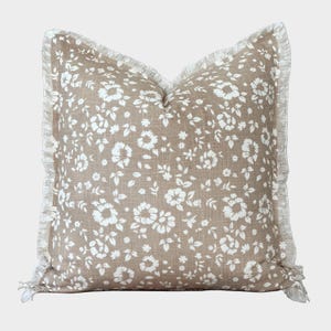 Op de afbeelding: Vierkant sierkussen met bloemenpatroon. Het kussen heeft een bruine achtergrond met witte bloemen en bladeren. De randen zijn afgewerkt met een witte franje. Dit decoratieve kussen is ideaal voor een bed of bank.