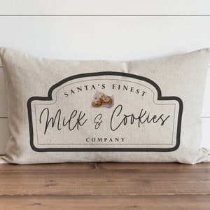 Pode incluir: Uma almofada bege com um gráfico preto e branco de um logotipo vintage que diz "Santa's Finest Milk & Cookies Company".