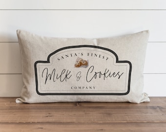 Puede incluir: Una almohada beige con un gráfico en blanco y negro de un logotipo vintage que dice "Santa's Finest Milk & Cookies Company".