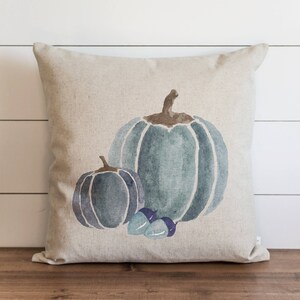 Op de afbeelding: Een decoratief kussen met een aquarelontwerp van twee blauwe pompoenen en eikels. Het kussen is gemaakt van een licht beige linnen stof.