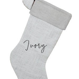 May include: Ivory Christmas stocking with a neutral-colored cuff and a white tassel. The word "Ivory" is written in a cursive font on the front of the stocking. The stocking is made of a textured fabric.