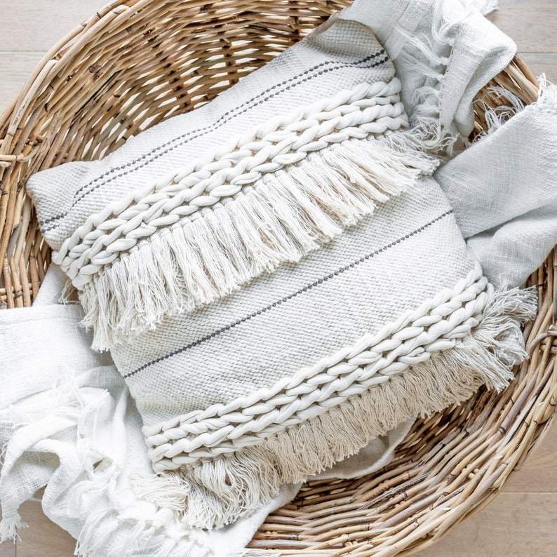 Puede incluir: Un coj&iacute;n decorativo blanco roto con dise&ntilde;o tejido y detalles de flecos. El coj&iacute;n tiene rayas grises oscuras y una textura trenzada. Descansa en una cesta de mimbre con una manta a juego.