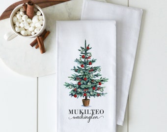 Peut inclure: Une serviette de cuisine blanche avec une illustration d'un sapin de Noël vert avec des décorations rouges et argentées. Le texte "MUKILTEO washington" est imprimé sous l'arbre.
