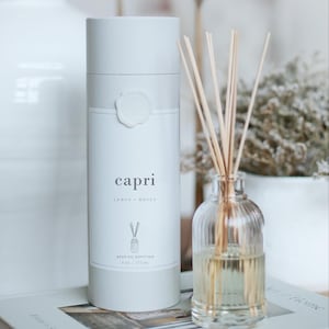 Könnte beinhalten: Ein weißer Capri-Schilföl-Diffusor mit einem Etikett mit der Aufschrift "Capri Lemon + Honey". Der Diffusor befindet sich neben einer klaren Glasflasche mit Schilf. Die Flasche enthält eine gelbe Flüssigkeit. Der Diffusor ist 177 ml.
