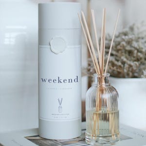 Puede incluir: Un juego de difusor de aceite de caña blanco con la palabra "weekend" impresa en la etiqueta. El juego incluye una botella de vidrio transparente con cañas y un recipiente cilíndrico. La etiqueta también incluye la fragancia "cuero + cardamomo" y el volumen, 177 ml.