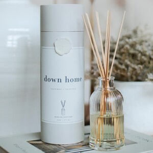 May include: A white reed oil diffuser with the text "down home" and "Saffron + Vetiver". A clear glass bottle with reeds is next to the diffuser. The diffuser is 6 oz / 177 ml.