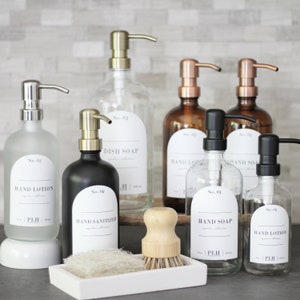 May include: A set of eight glass bottles with pumps, labeled with numbers and product names, including hand lotion, hand sanitizer, dish soap, and hand soap. The bottles are arranged on a counter with a wooden brush and a white tray.