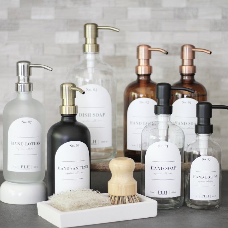 May include: A set of eight glass bottles with pumps, labeled with numbers and product names, including hand lotion, hand sanitizer, dish soap, and hand soap. The bottles are arranged on a counter with a wooden brush and a white tray.