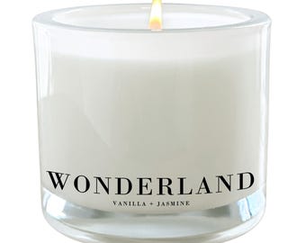 Puede incluir: Un tarro de vela de vidrio transparente con una vela blanca en su interior. La vela está encendida y la llama es visible. El tarro tiene una etiqueta negra con el texto "WONDERLAND" y "VANILLA + JASMINE" impreso en ella.