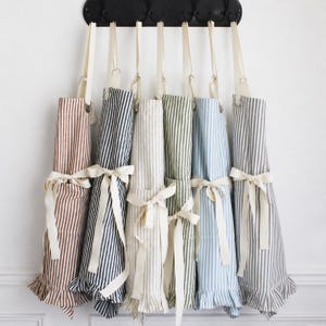 May include: A collection of striped aprons in various colors, including brown, navy, and blue, hanging on a black rack. Each apron features a ruffled hem and a cream-colored tie at the waist. The aprons are made of a soft fabric.