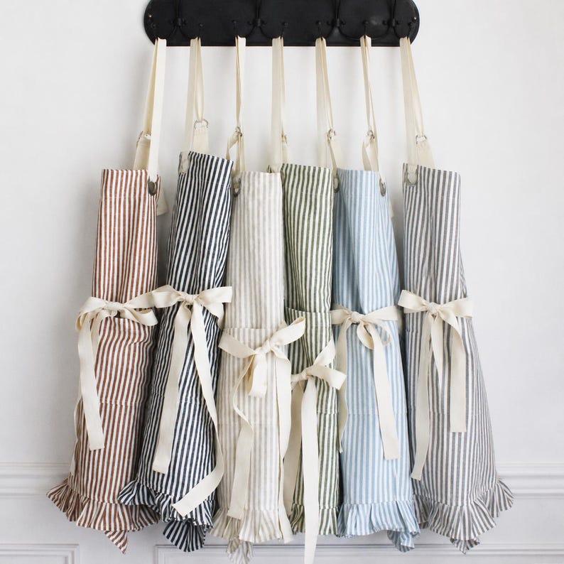 May include: A collection of striped aprons in various colors, including brown, navy, and blue, hanging on a black rack. Each apron features a ruffled hem and a cream-colored tie at the waist. The aprons are made of a soft fabric.
