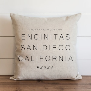 Puede incluir: Un cojín cuadrado de lino beige con texto impreso en negro. El texto dice there's no place like home, seguido de ENCINITAS, SAN DIEGO, CALIFORNIA y el código postal 92024 en una fuente sans-serif clara centrada en la tela.