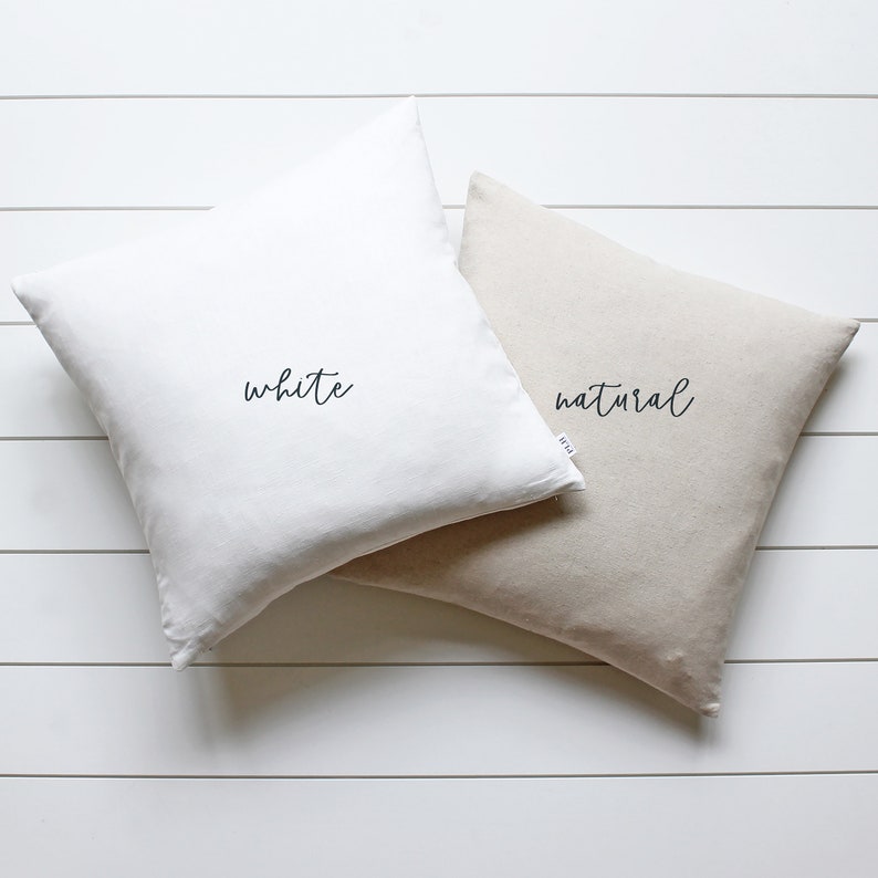 Puede incluir: Dos almohadas cuadradas, una blanca y otra de lino natural, con las palabras "white" y "natural" escritas en cursiva negra en cada almohada.