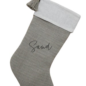 May include: A gray Christmas stocking with a white cuff and a gray tassel. The word "Sand" is written in a cursive font on the front of the stocking. A loop is attached for hanging.