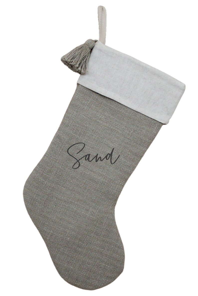 May include: A gray Christmas stocking with a white cuff and a gray tassel. The word "Sand" is written in a cursive font on the front of the stocking. A loop is attached for hanging.