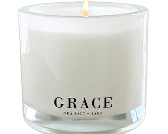 Puede incluir: Un tarro de vela de vidrio transparente con una vela blanca en su interior. La vela está encendida y la llama es visible. El tarro tiene una etiqueta negra que dice "GRACE SEA SALT + SAGE".