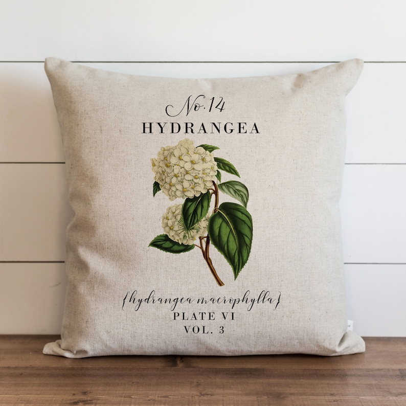 May include: A beige linen pillow with a botanical illustration of a white hydrangea flower with green leaves. The pillow has the text "No. 14 HYDRANGEA {hydrangea macrophylla} PLATE VI VOL. 3" printed on it.