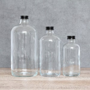 May include: Three clear glass bottles with black lids. The bottles are different sizes and are arranged in a row from largest to smallest.
